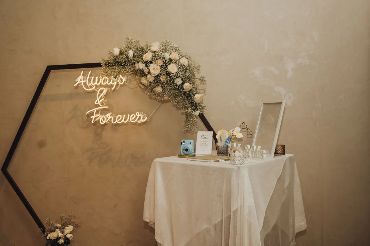 Gästebuch-Tisch mit Sofortbildkamera und Hinweiskarte vor einem Blumenbogen mit Neon-Schriftzug „Always & Forever“